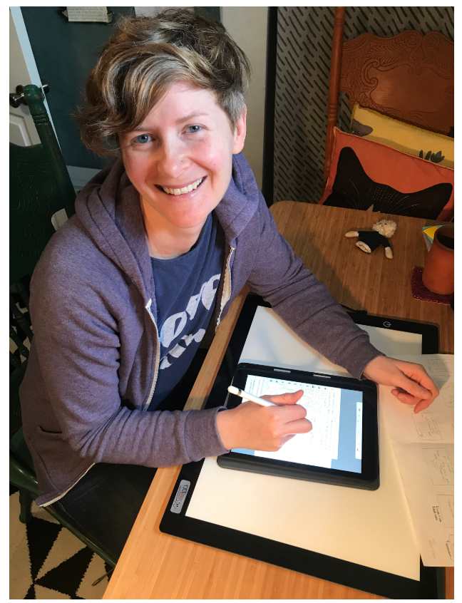Photo of scientist and comic artist Cara Gomally sitting and looking at the camera and smiling. She is at a desk with some drawings and her ipad, working on her comics.