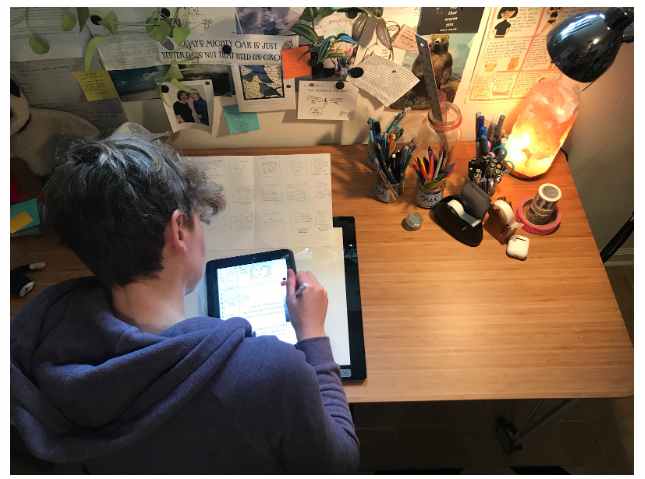 Photo of scientist and comic artist Cara Gomally sitting at her desk with her drawings and her ipad, working on her comics. Photo taken from birds eye view.