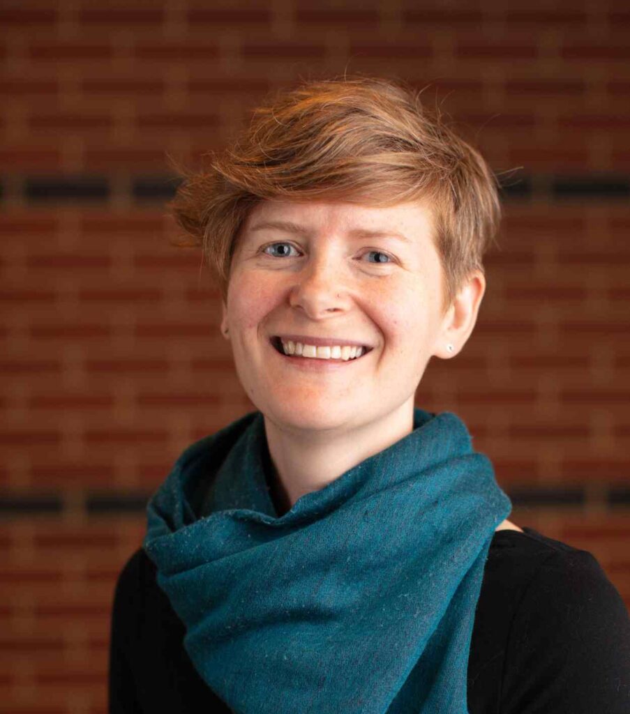 A cheerful person with short hair and a teal scarf smiles warmly against a red brick wall background.