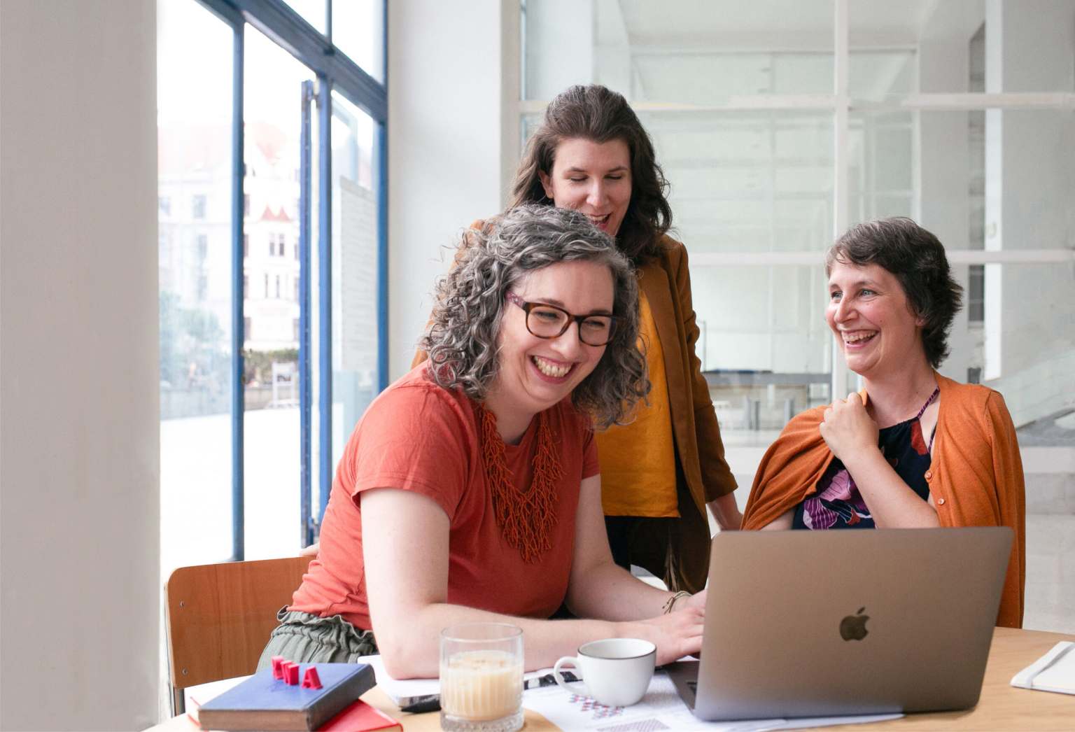 Three women laugh and converse around a laptop in a brightly lit cafe with books and coffee on the table, indicating a casual meeting or friendly gathering.